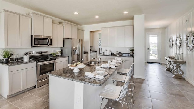 kitchen featuring appliances with stainless steel finishes, a kitchen breakfast bar, light tile patterned floors, dark stone countertops, and an island with sink