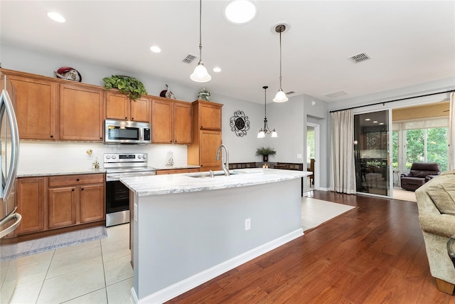 kitchen with light wood-type flooring, light stone counters, stainless steel appliances, a kitchen island with sink, and decorative light fixtures