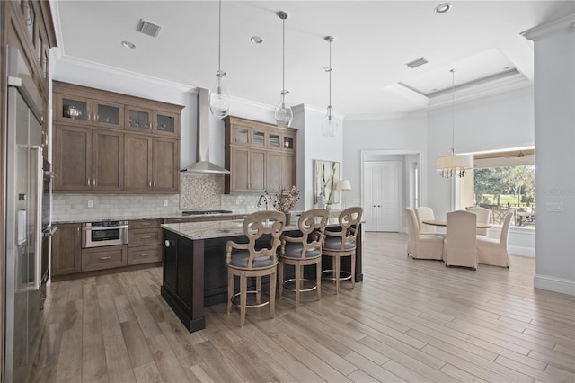 kitchen with pendant lighting, light wood-type flooring, a center island with sink, and wall chimney range hood