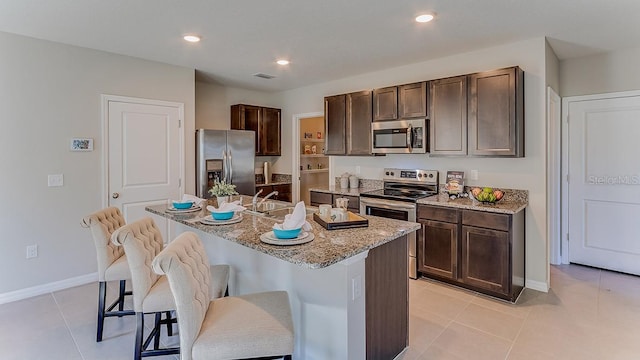 kitchen with dark brown cabinetry, light stone counters, a kitchen bar, a center island with sink, and appliances with stainless steel finishes