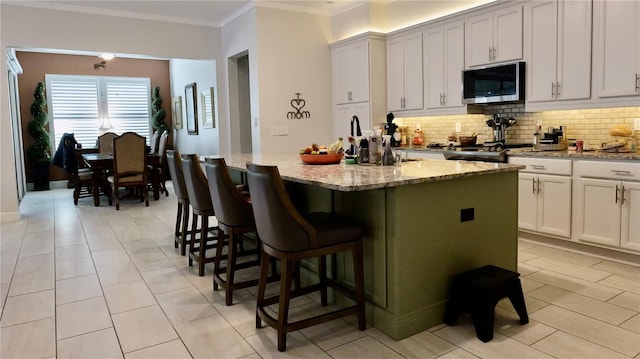 kitchen with light stone countertops, decorative backsplash, crown molding, a center island with sink, and white cabinetry