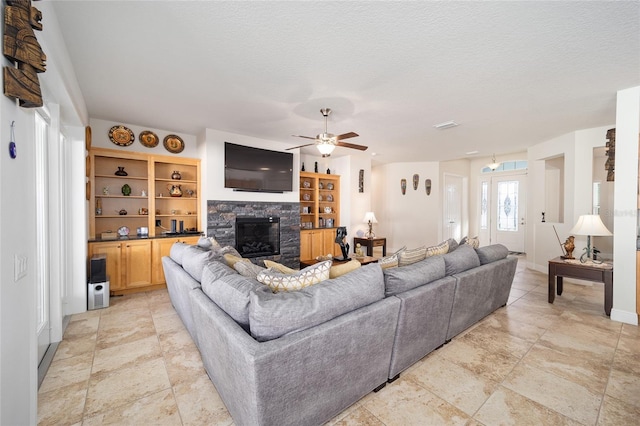 living room with ceiling fan, a stone fireplace, built in features, and a textured ceiling