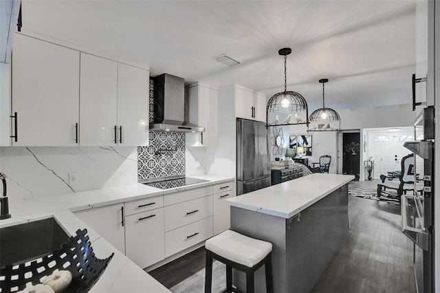 kitchen with black electric stovetop, hanging light fixtures, white cabinets, wall chimney range hood, and stainless steel refrigerator