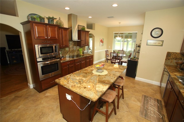 kitchen with a center island, wall chimney exhaust hood, light stone counters, a kitchen bar, and stainless steel appliances