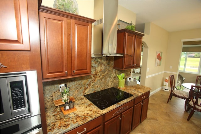 kitchen with light stone countertops, decorative backsplash, wall chimney exhaust hood, black electric cooktop, and stainless steel microwave