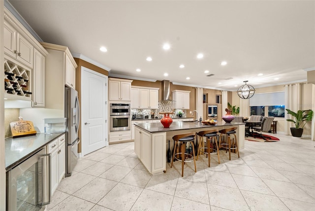 kitchen with wall chimney range hood, wine cooler, an island with sink, cream cabinetry, and appliances with stainless steel finishes