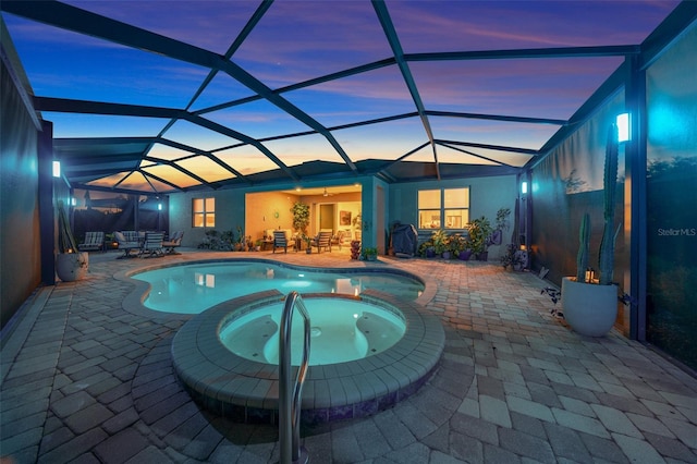 pool at dusk with an in ground hot tub, glass enclosure, and a patio area