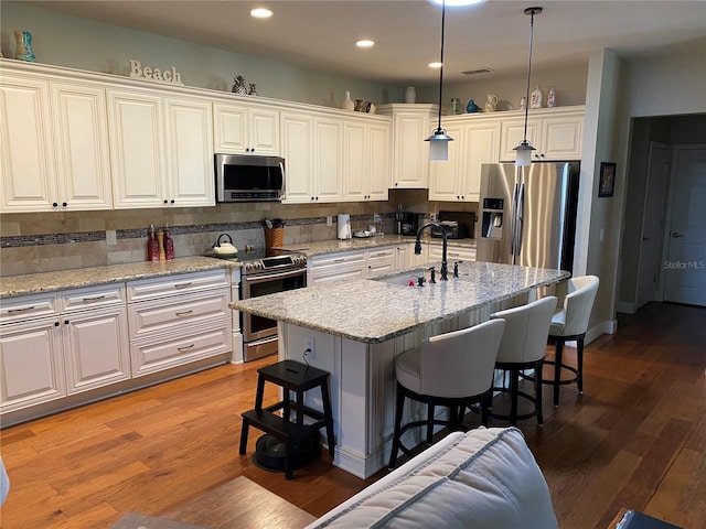 kitchen with sink, stainless steel appliances, hardwood / wood-style floors, decorative light fixtures, and a center island with sink