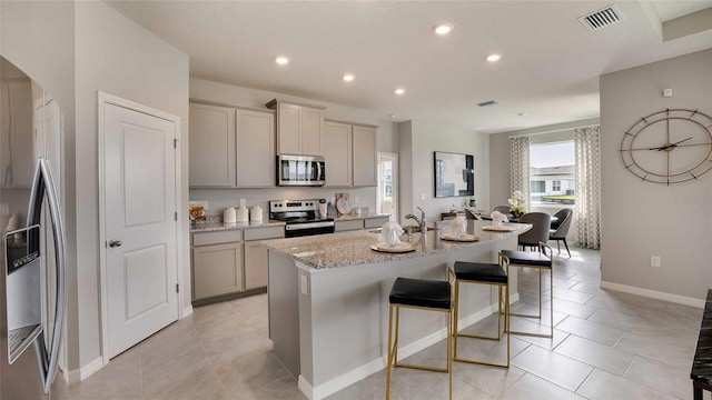 kitchen featuring light stone countertops, a kitchen breakfast bar, gray cabinetry, stainless steel appliances, and a kitchen island with sink