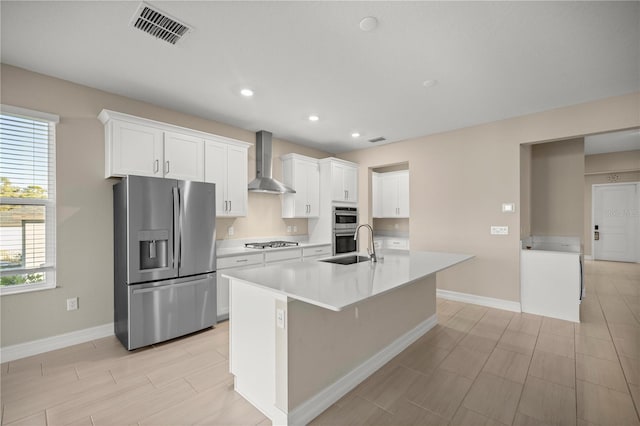 kitchen with sink, wall chimney exhaust hood, stainless steel appliances, an island with sink, and white cabinets