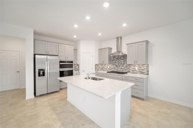 kitchen with sink, wall chimney exhaust hood, light stone counters, a kitchen island with sink, and appliances with stainless steel finishes