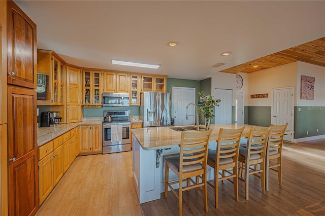 kitchen with a center island with sink, sink, light stone countertops, light wood-type flooring, and appliances with stainless steel finishes