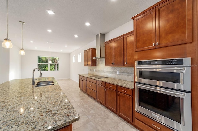 kitchen featuring sink, stainless steel double oven, wall chimney range hood, light stone counters, and pendant lighting