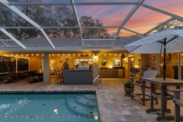 pool at dusk with glass enclosure, a patio area, a bar, and an outdoor kitchen