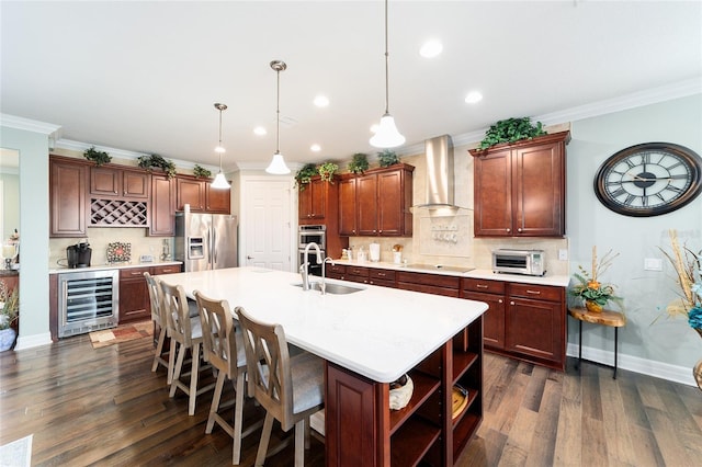 kitchen featuring a center island with sink, wall chimney range hood, decorative light fixtures, stainless steel appliances, and beverage cooler