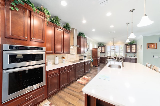 kitchen featuring stainless steel double oven, decorative light fixtures, wall chimney exhaust hood, and sink