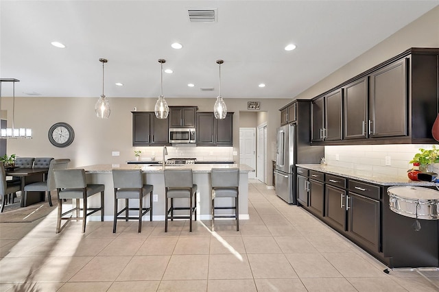 kitchen with appliances with stainless steel finishes, light stone counters, dark brown cabinets, a kitchen island with sink, and hanging light fixtures