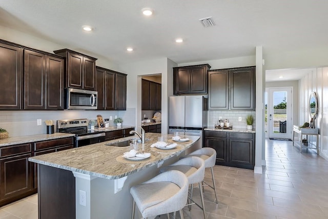 kitchen with appliances with stainless steel finishes, light stone counters, dark brown cabinets, a kitchen island with sink, and sink