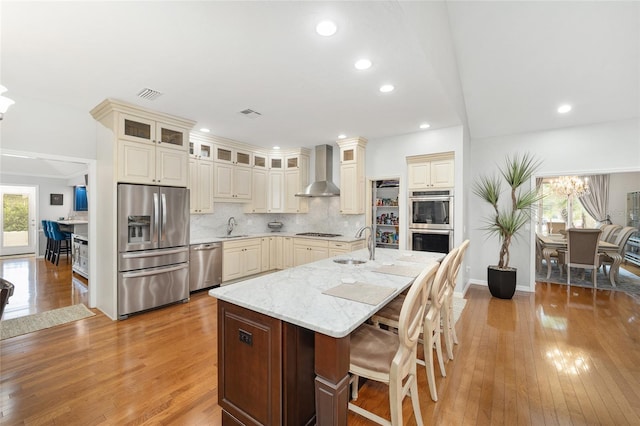 kitchen with stainless steel appliances, a kitchen island with sink, wall chimney exhaust hood, and light hardwood / wood-style floors