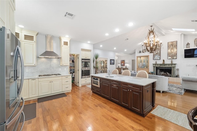 kitchen with wall chimney exhaust hood, stainless steel appliances, lofted ceiling with skylight, a center island with sink, and light hardwood / wood-style floors