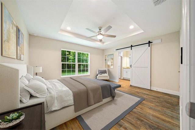 bedroom with a raised ceiling, ceiling fan, a barn door, connected bathroom, and wood-type flooring