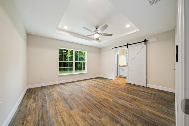 unfurnished bedroom with a tray ceiling, a barn door, ceiling fan, and dark hardwood / wood-style flooring