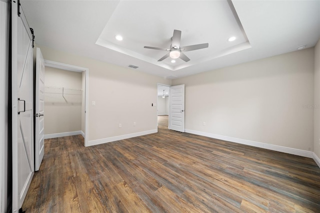 unfurnished bedroom with a raised ceiling, a barn door, ceiling fan, and dark hardwood / wood-style flooring