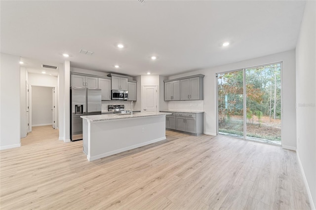 kitchen featuring gray cabinetry, backsplash, an island with sink, appliances with stainless steel finishes, and light wood-type flooring