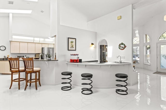 kitchen with kitchen peninsula, stainless steel fridge, a breakfast bar, light brown cabinets, and high vaulted ceiling