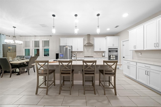 kitchen featuring wall chimney exhaust hood, decorative light fixtures, a kitchen bar, a kitchen island with sink, and appliances with stainless steel finishes