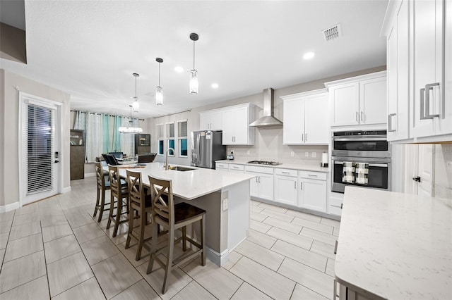 kitchen with white cabinets, wall chimney exhaust hood, a center island with sink, and stainless steel appliances