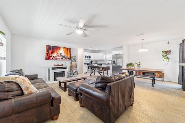 living room with ceiling fan with notable chandelier, a stone fireplace, a textured ceiling, and light hardwood / wood-style flooring