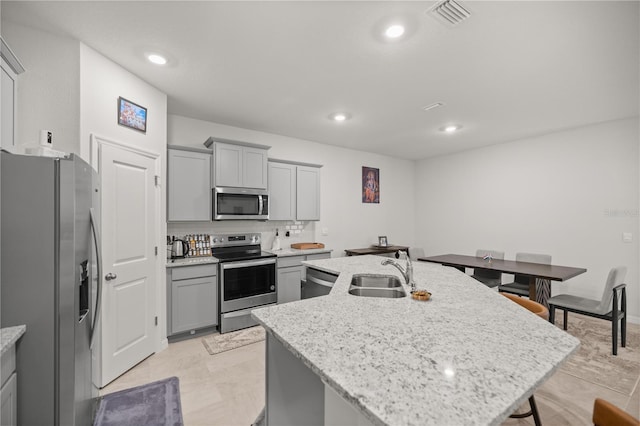 kitchen featuring a kitchen island with sink, sink, gray cabinets, light stone counters, and stainless steel appliances