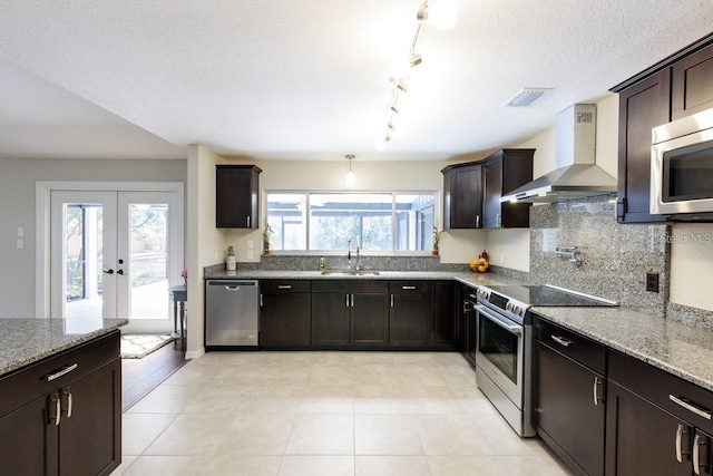 kitchen with appliances with stainless steel finishes, french doors, dark brown cabinetry, sink, and wall chimney range hood