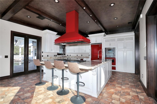 kitchen with stainless steel microwave, a kitchen island with sink, white cabinets, french doors, and island range hood