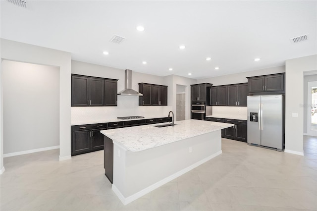 kitchen featuring light stone countertops, sink, stainless steel appliances, wall chimney range hood, and an island with sink