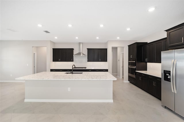 kitchen with a kitchen island with sink, wall chimney range hood, sink, light stone countertops, and stainless steel fridge with ice dispenser