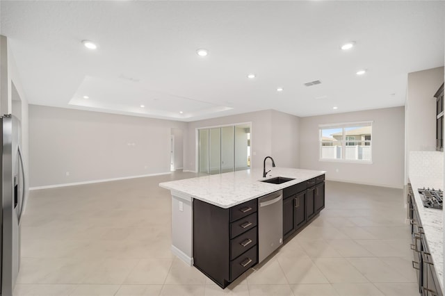 kitchen with sink, stainless steel appliances, a raised ceiling, light stone counters, and a kitchen island with sink