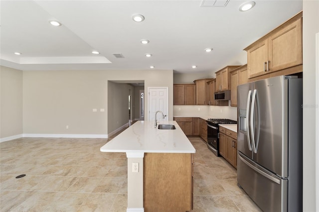 kitchen with appliances with stainless steel finishes, light stone counters, a raised ceiling, sink, and an island with sink