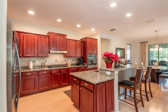 kitchen with appliances with stainless steel finishes, light stone counters, an inviting chandelier, hanging light fixtures, and an island with sink