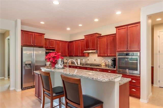 kitchen with a breakfast bar area, decorative backsplash, light stone counters, stainless steel appliances, and a center island with sink