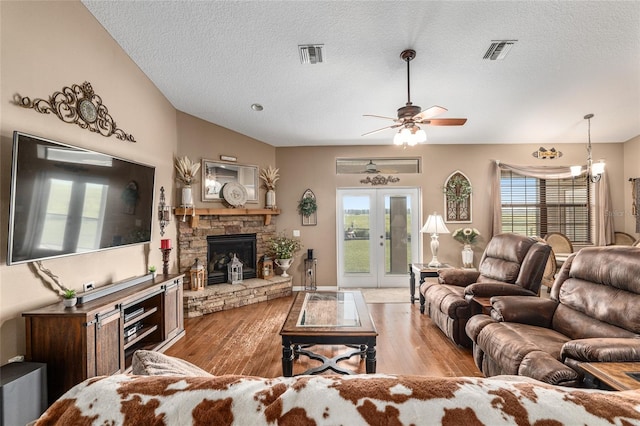 living room featuring french doors, light wood-type flooring, ceiling fan with notable chandelier, a textured ceiling, and a stone fireplace