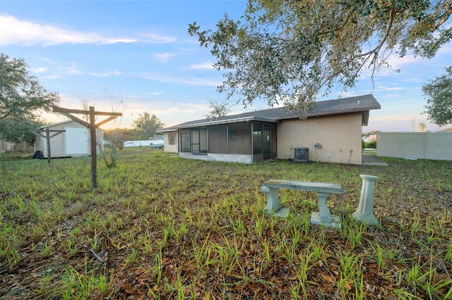 back house at dusk with central air condition unit, a lawn, a storage shed, and a sunroom