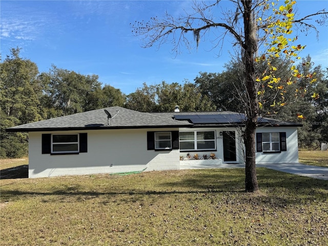 ranch-style home featuring concrete block siding, roof with shingles, roof mounted solar panels, and a front lawn