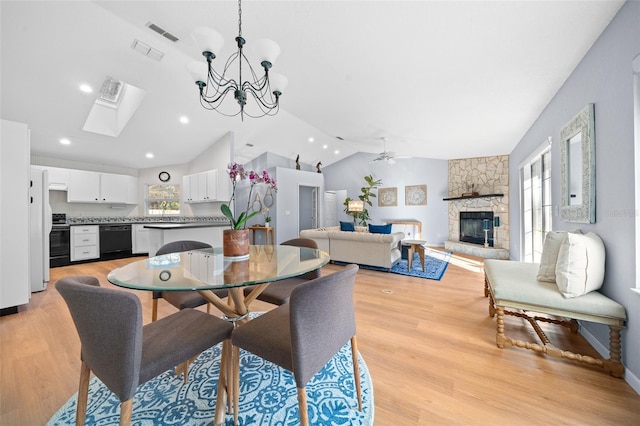 dining space featuring light wood-type flooring, ceiling fan with notable chandelier, a stone fireplace, and lofted ceiling with skylight