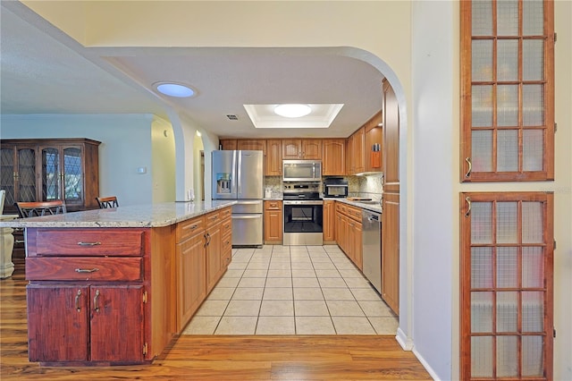 kitchen with light stone countertops, stainless steel appliances, tasteful backsplash, a tray ceiling, and light wood-type flooring