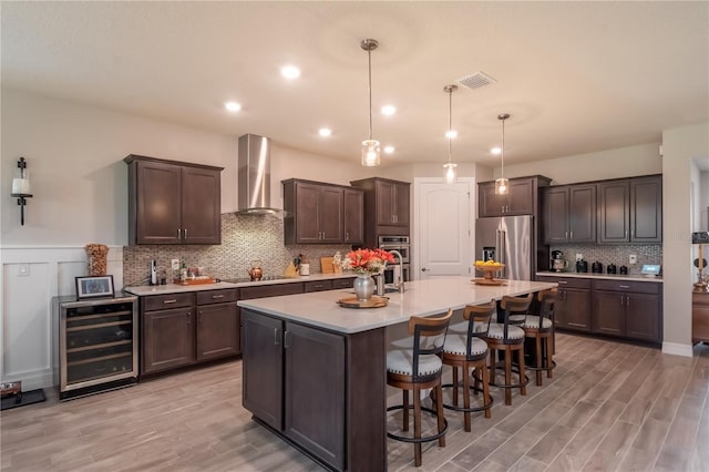 kitchen with decorative light fixtures, wall chimney exhaust hood, dark brown cabinets, stainless steel appliances, and beverage cooler