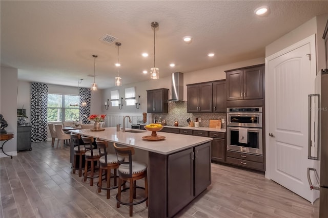 kitchen with pendant lighting, a kitchen island with sink, wall chimney exhaust hood, appliances with stainless steel finishes, and dark brown cabinetry