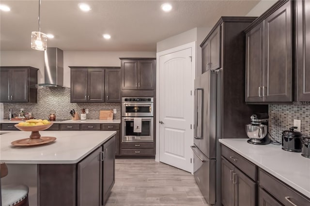 kitchen featuring appliances with stainless steel finishes, backsplash, dark brown cabinetry, wall chimney range hood, and hanging light fixtures