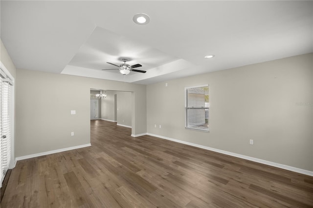 empty room with dark hardwood / wood-style floors, plenty of natural light, ceiling fan with notable chandelier, and a tray ceiling